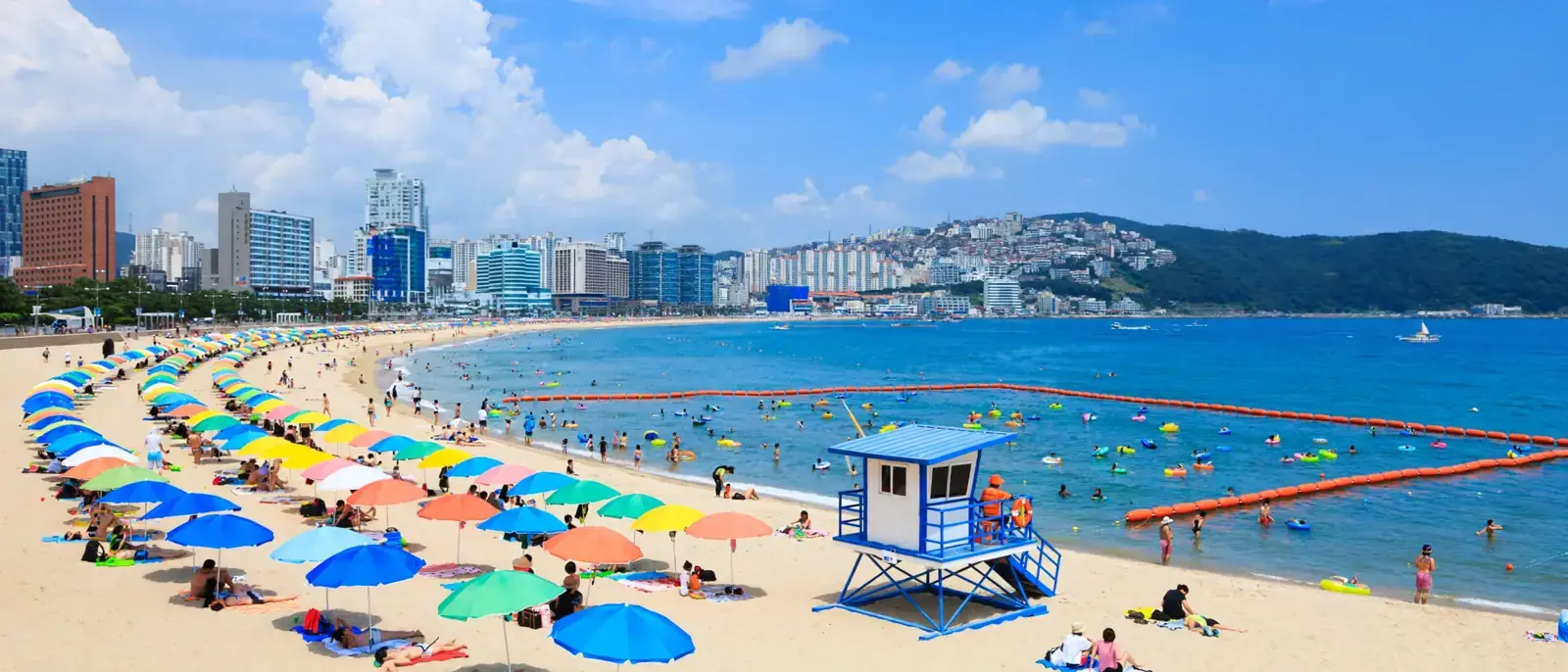When to visit Korean beaches in peak summer — rows of colorful parasols, swimmers in a buoyed swim zone and a lifeguard tower