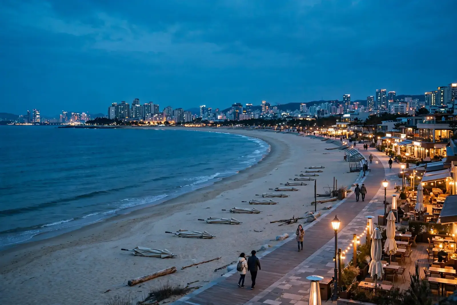 A South Korea Travel Tips for a Beach Trip promenade at twilight, with folded parasols, cafe lights and a couple strolling beside the sea